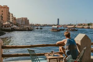 Man sitting at a waterfront café watching small boats pass along a busy urban waterway on a sunny day.