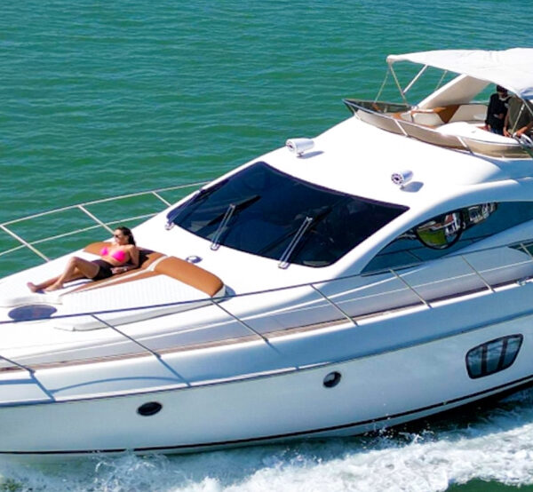 A white 60 ft Azimut “Shedai” yacht cruising on turquoise water, with a woman sunbathing on the bow and guests relaxing on the flybridge under a canopy.